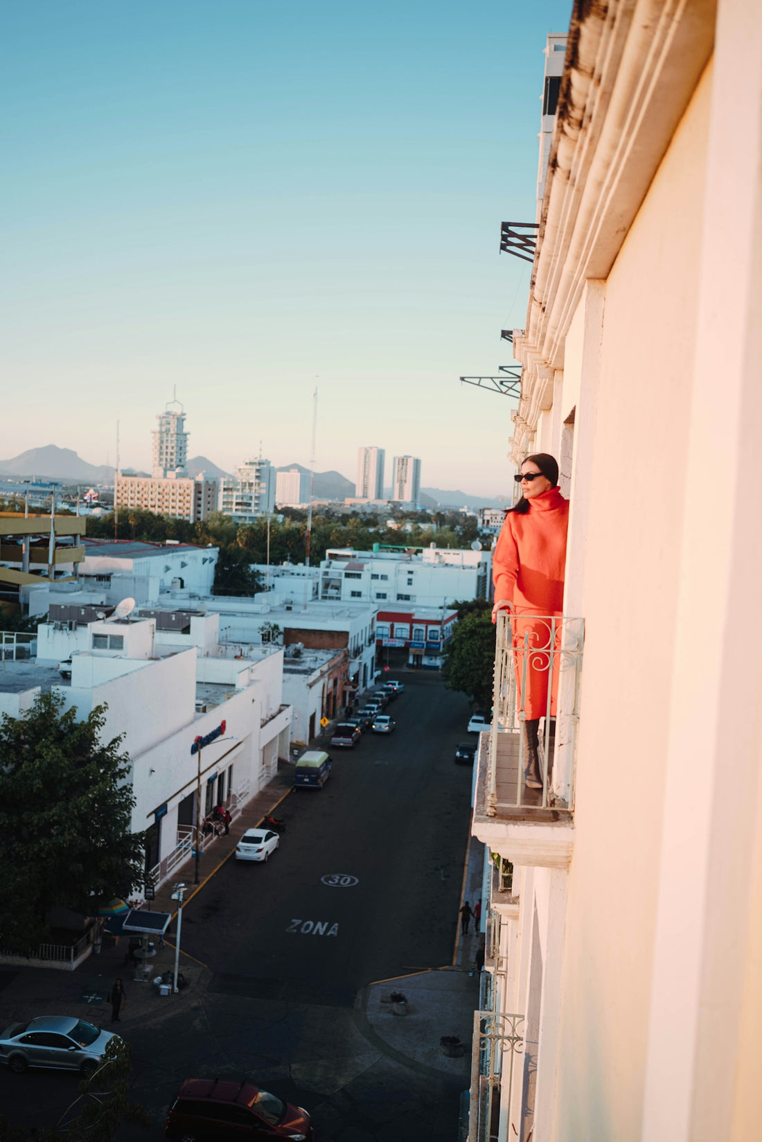 Woman in orange coat on balcony overlooking city street.